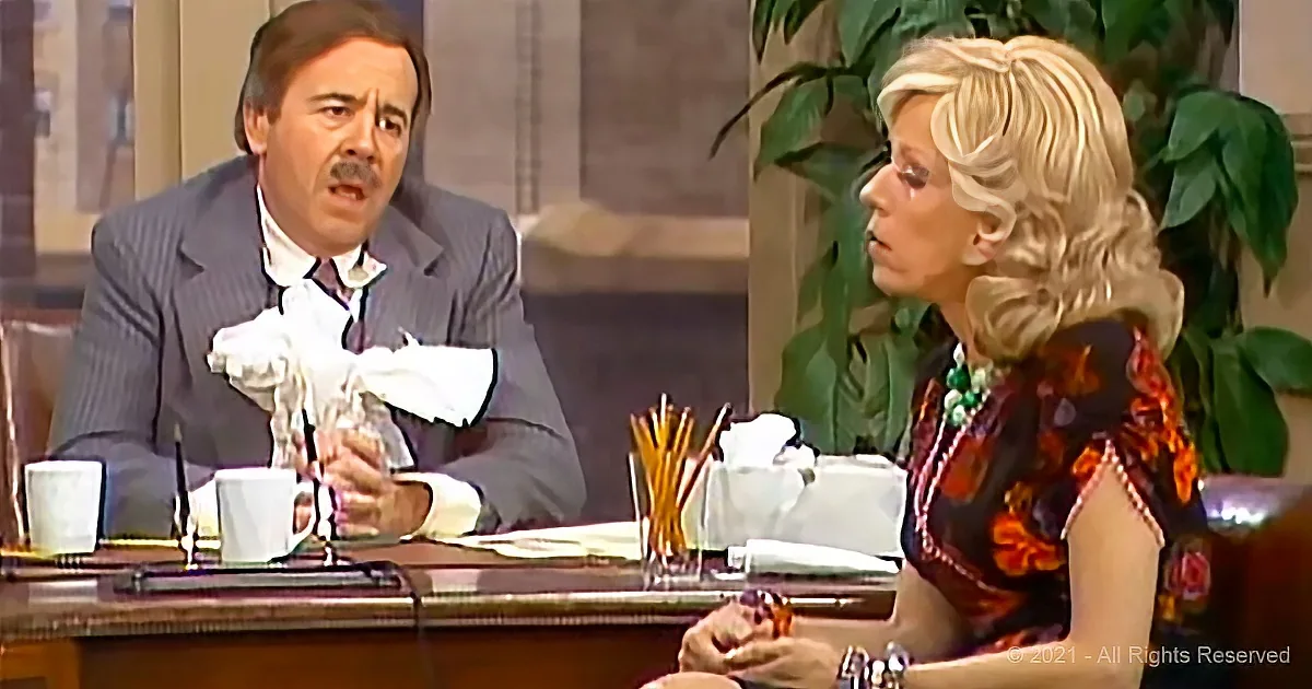 A man in a suit holds a crumpled paper while a woman in a floral dress looks on, both seated at a table with office supplies.