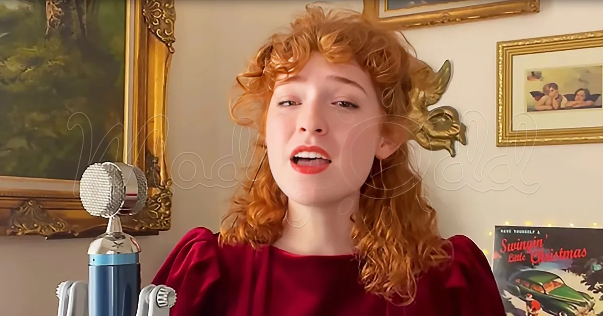 A person with curly red hair sings into a microphone, wearing a red top, in a room with framed pictures and a Christmas...