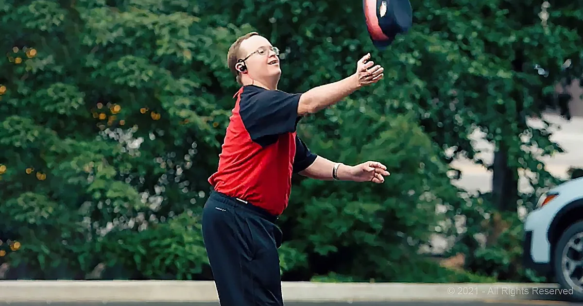 A man in a red and black shirt plays with a ball in a park with trees and a car in the background.