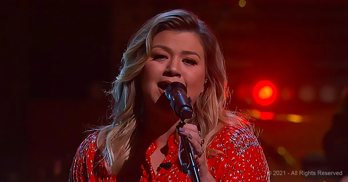 A woman in a red patterned dress sings into a microphone on stage with red lighting.