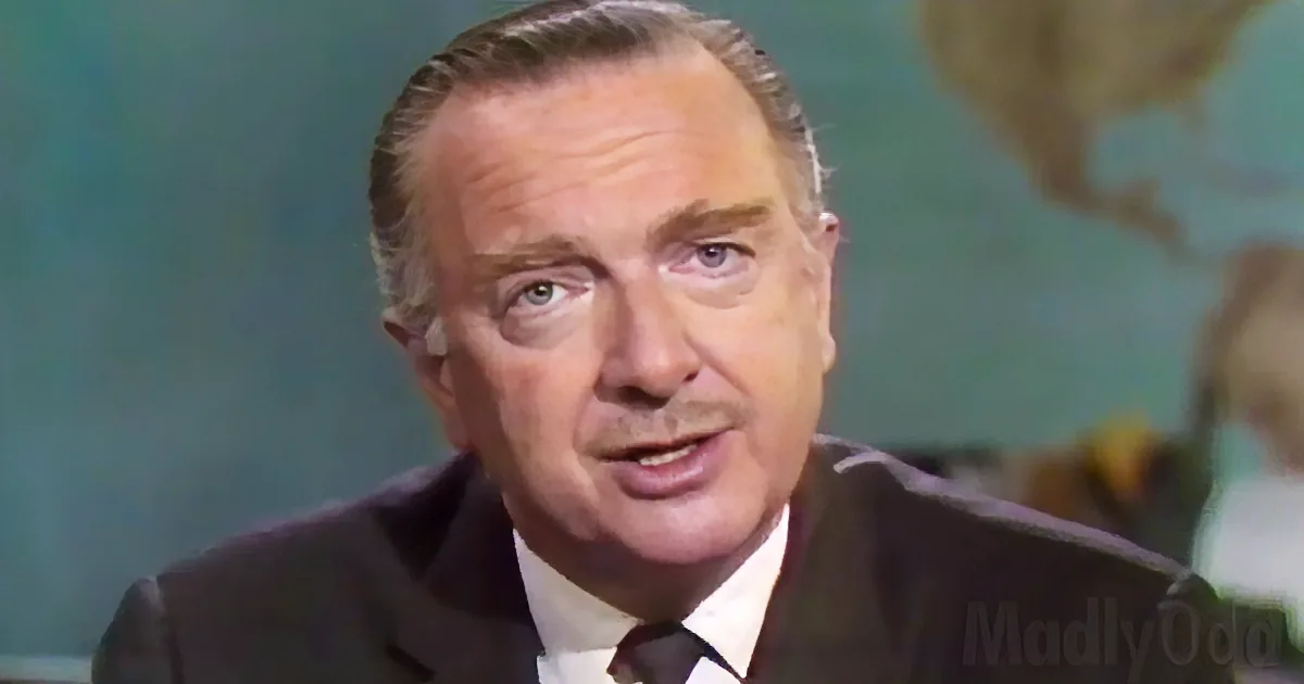 A man in a suit and tie is speaking, with a map in the background.