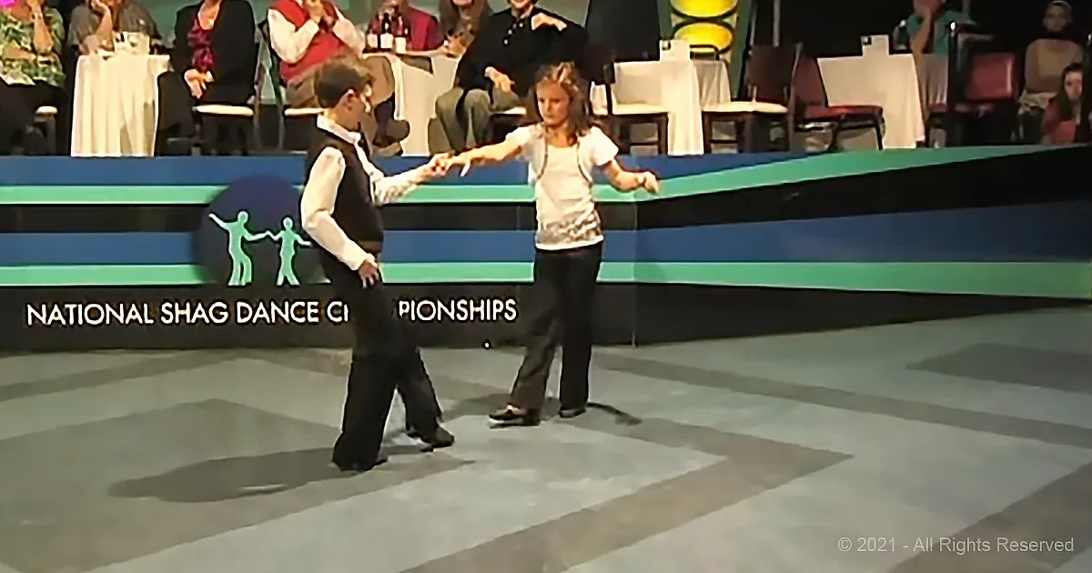 A boy and girl dance in a Shag Dance Championship, holding hands and moving gracefully on stage.