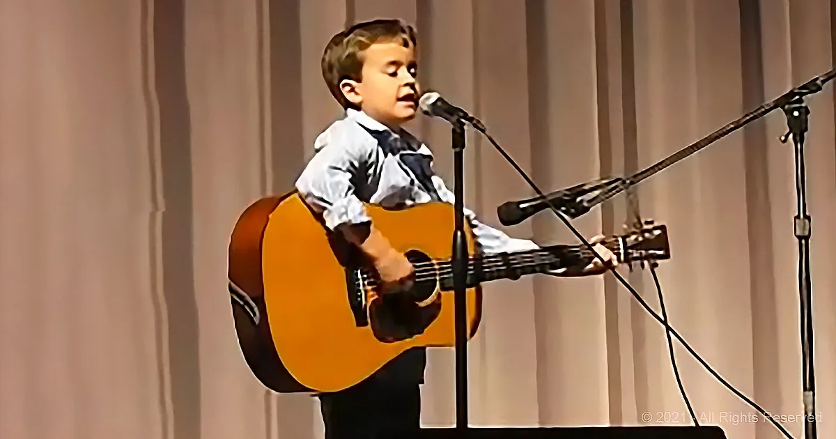 A young boy sings into a microphone while playing an acoustic guitar on stage.