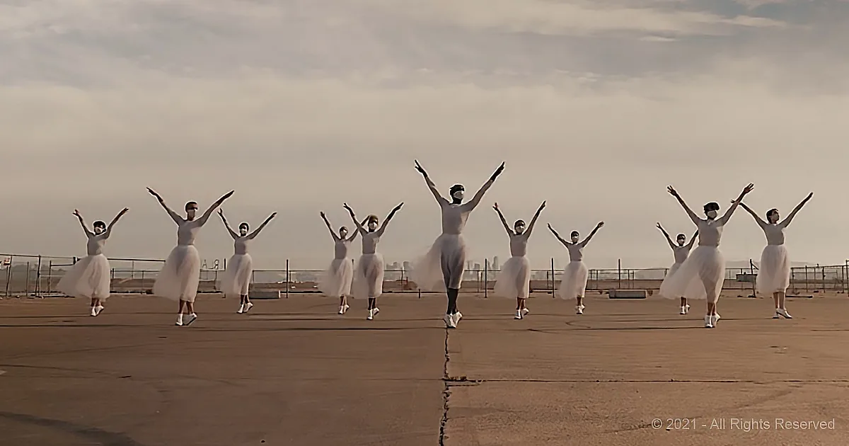 Dancers in white tutus perform synchronized arm movements on a rooftop under a cloudy sky.