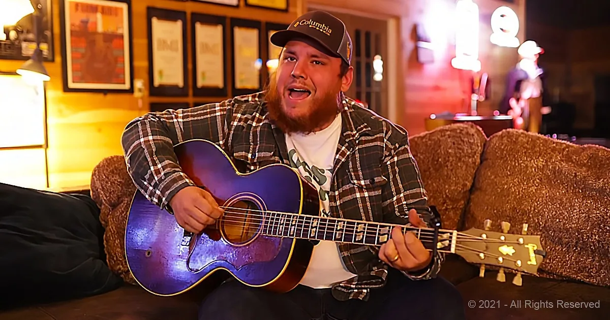 A man in a plaid shirt and cap plays an acoustic guitar on a couch in a warmly lit room.