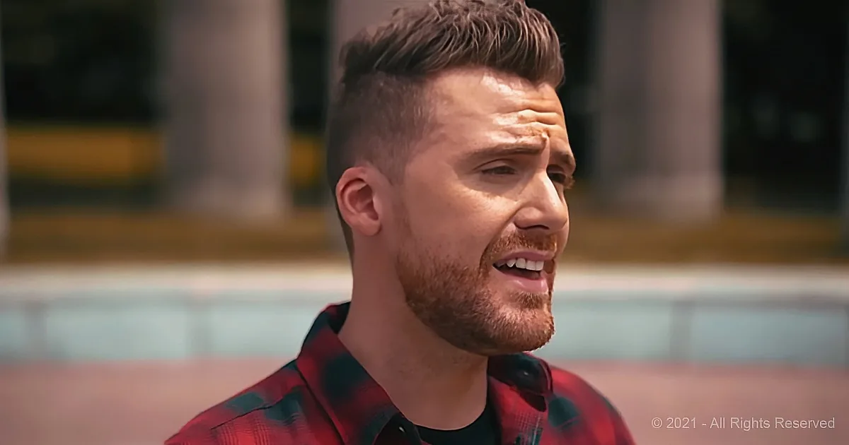 A man with short hair and a beard, wearing a red plaid shirt, is looking to the side with a slight smile.