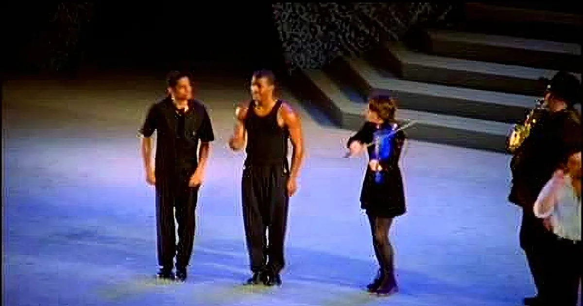 Five dancers in white shirts and black pants perform synchronized steps on a stage with a cityscape backdrop.