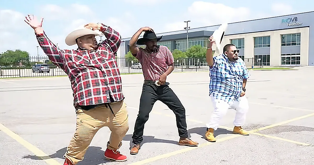 Three men in cowboy hats and casual attire dance in a parking lot, with a building in the background.