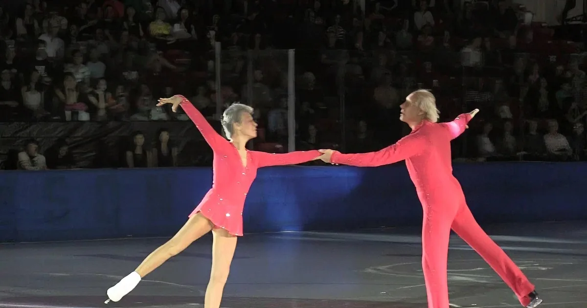 Two figure skaters in red costumes perform a graceful pose on ice, with one skater's leg extended.