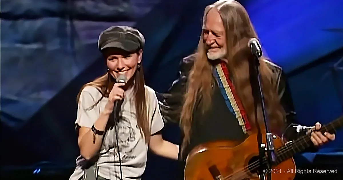 A woman sings into a microphone, smiling, while a man with long hair plays guitar behind her.