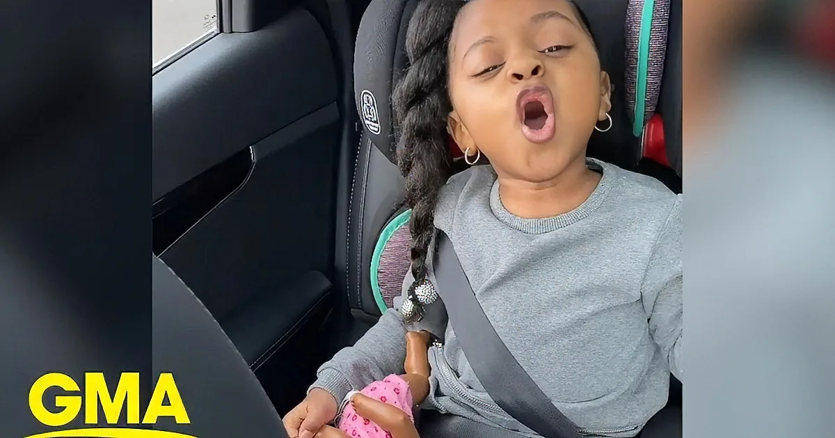 A young girl with braided hair sits in a car seat, wearing a light blue shirt and earrings, with her hand near her mouth.