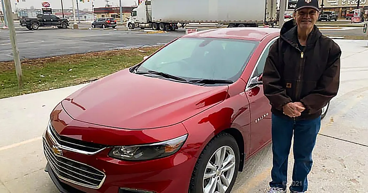 A man stands next to a red Chevrolet Malibu parked on a sidewalk near a road with traffic.