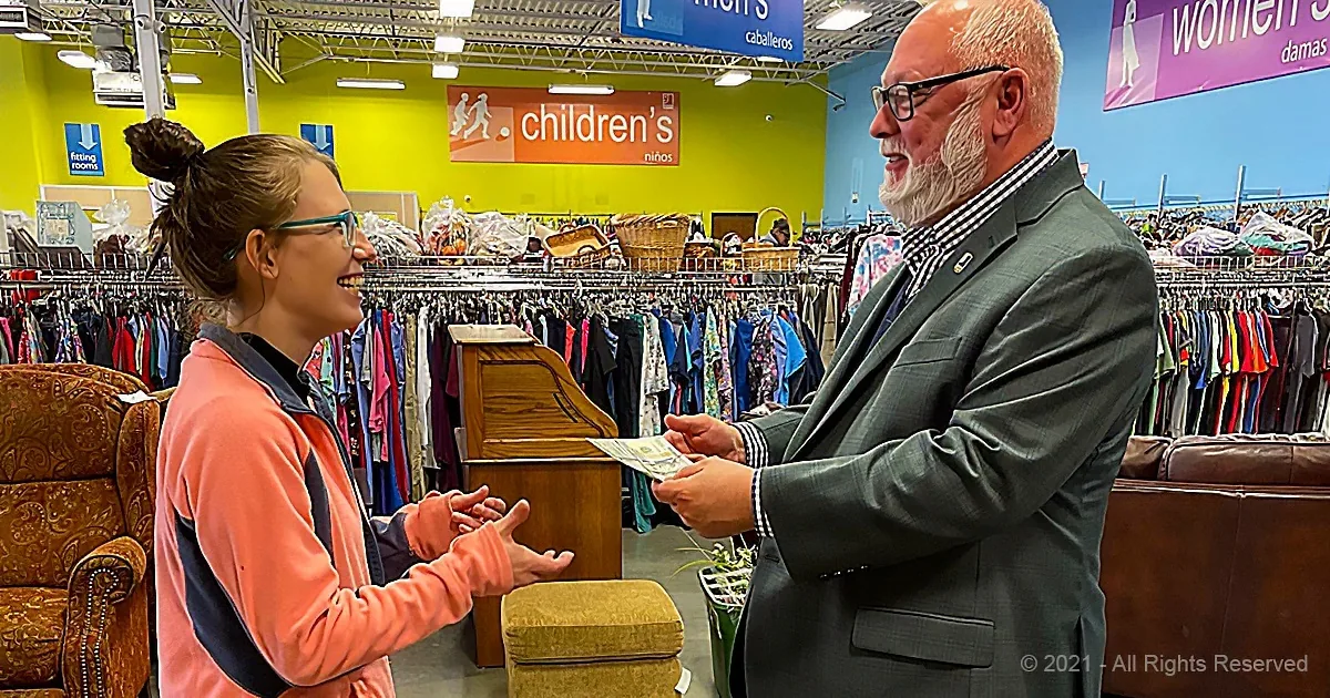 A man in a suit hands money to a woman in a thrift store, surrounded by racks of clothes.