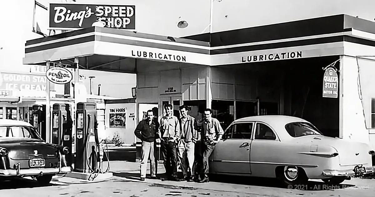 Four men stand outside Bing's Speed Shop, a vintage gas station with classic cars and service signs.
