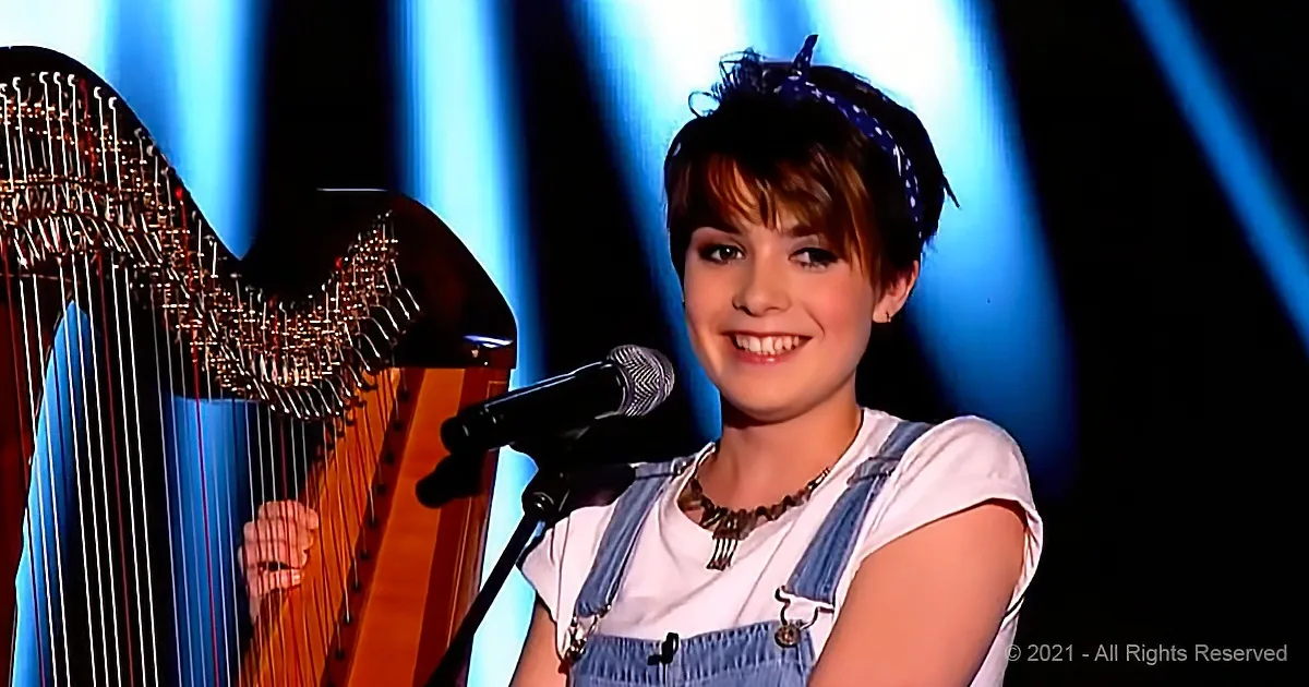 A smiling woman in denim overalls plays a harp on stage with a microphone in front of her.