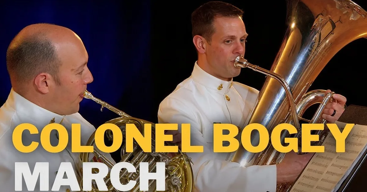 Two men in white uniforms play brass instruments on stage, one with a trombone, the other with a trumpet.