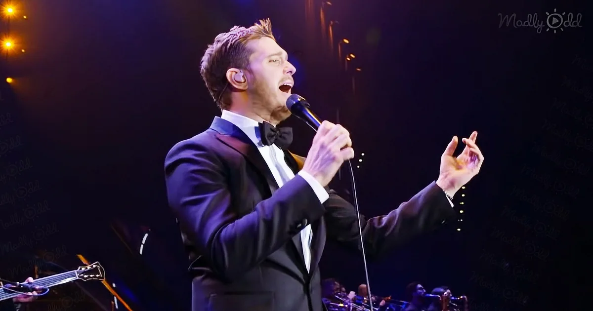 A man in a tuxedo sings passionately into a microphone on stage, surrounded by musicians.