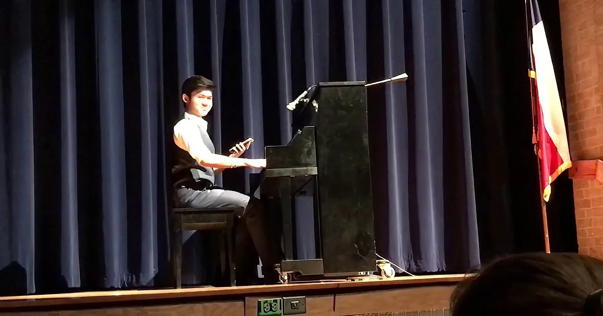 A person in a white shirt and gray vest plays a piano on stage with a microphone and blue curtains.