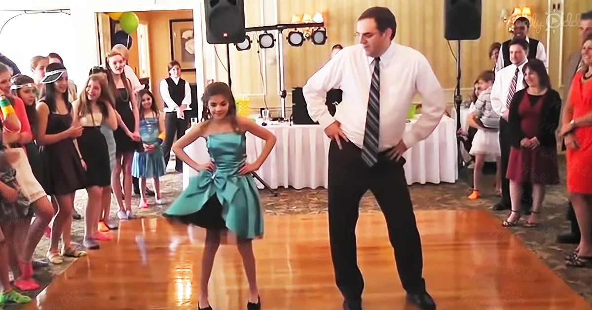 A girl in a teal dress dances with a man in a white shirt and tie on a wooden floor, surrounded by onlookers.