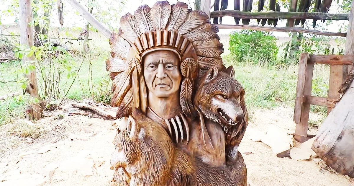 A carved wooden figure of an Indian chief with a wolf, wearing a feathered headdress, stands outdoors.