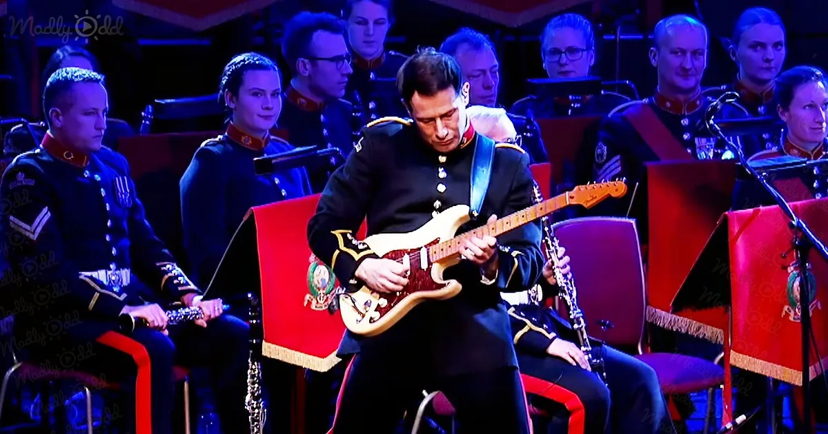 A man in military attire plays a guitar on stage, surrounded by musicians in uniform.