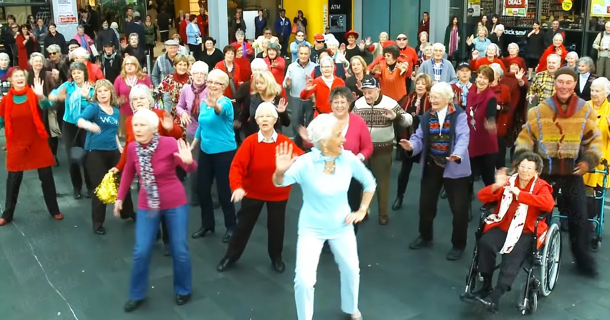 A group of elderly people, including a woman in a wheelchair, are dancing and waving in a public space.