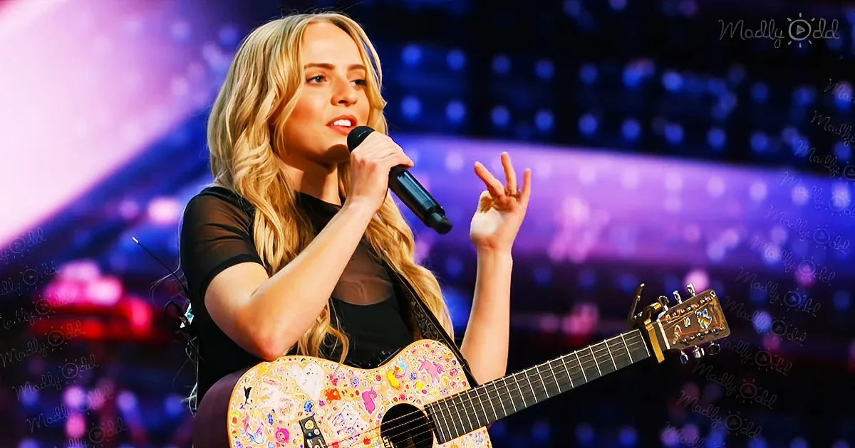 A woman with long blonde hair sings into a microphone while holding a decorated guitar on stage.
