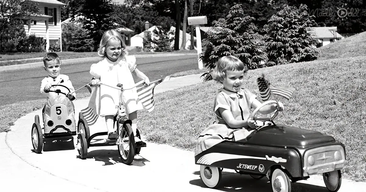 Three children ride vintage pedal cars on a suburban sidewalk, enjoying a sunny day.
