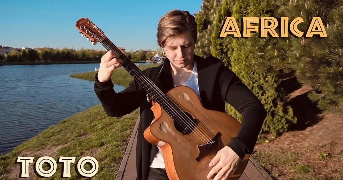 A person plays an acoustic guitar by a lake with buildings in the background.