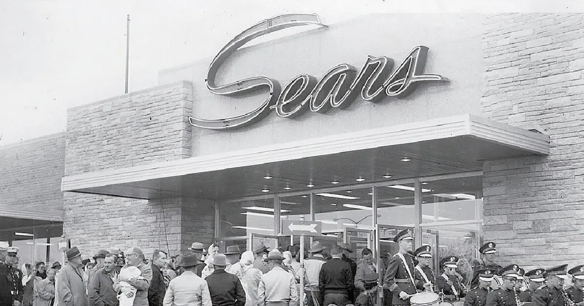 A vintage Sears store with palm trees, classic cars, and an American flag flying.