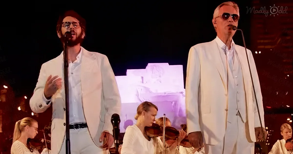 Two men in white suits sing on stage, accompanied by a string quartet and a projected image of a building.
