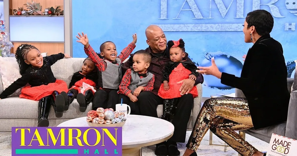 A man in a black suit sits with five children in festive attire, surrounded by Christmas decorations.