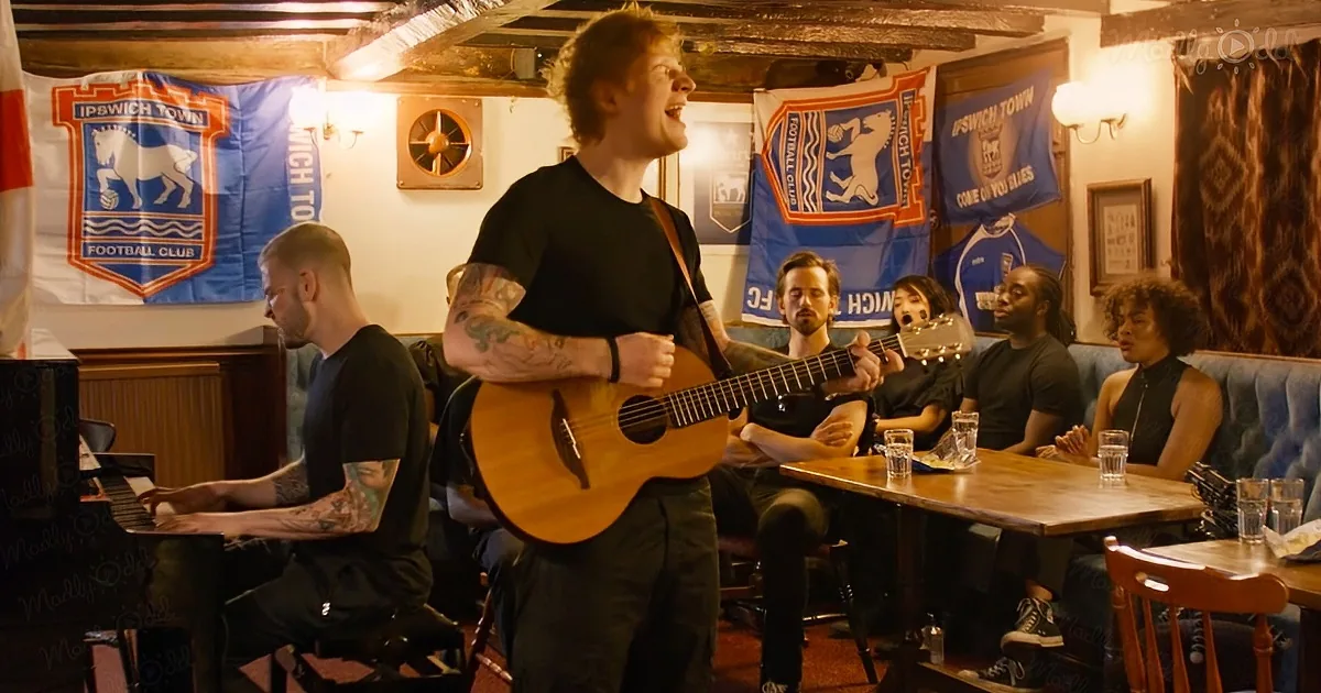 A man with tattoos plays guitar and sings in a pub with people watching and drinking.