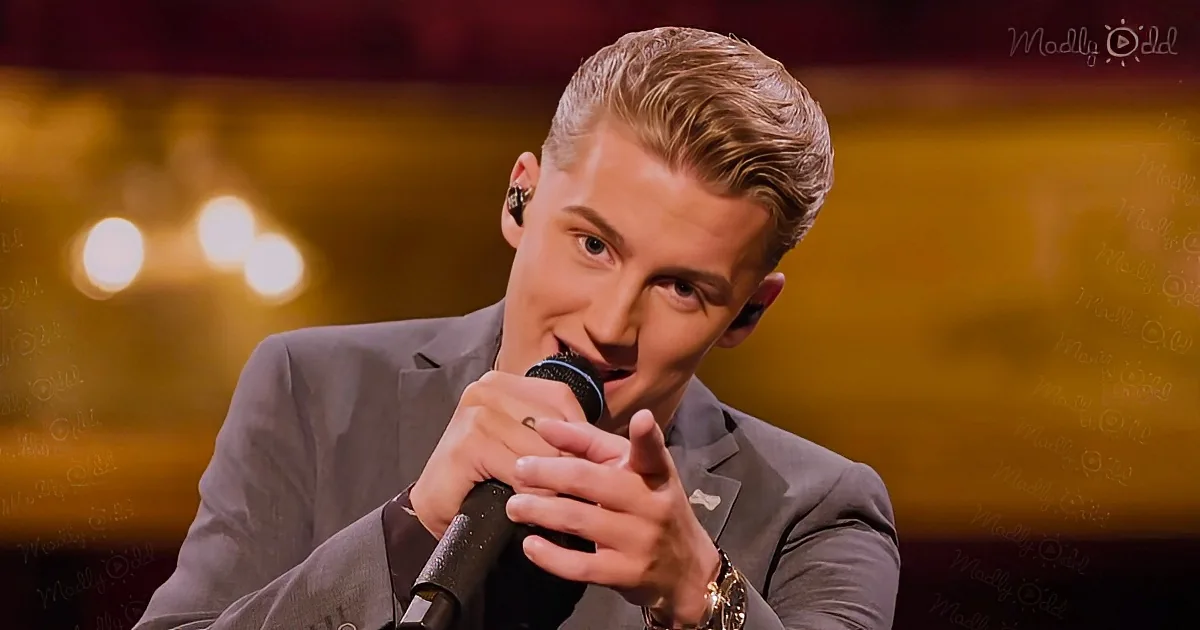 A man in a suit holds a microphone, wearing earbuds, and appears to be singing or speaking on stage.