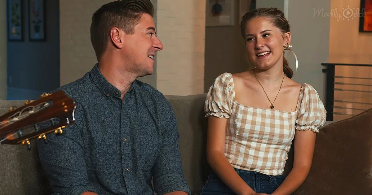 A man and woman are sitting on a couch, smiling and laughing together, with a guitar partially visible on the left.
