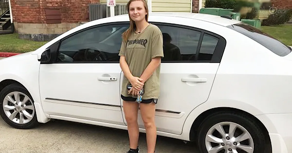 A person stands next to a white car, wearing a green t-shirt and black shorts, holding a drink.