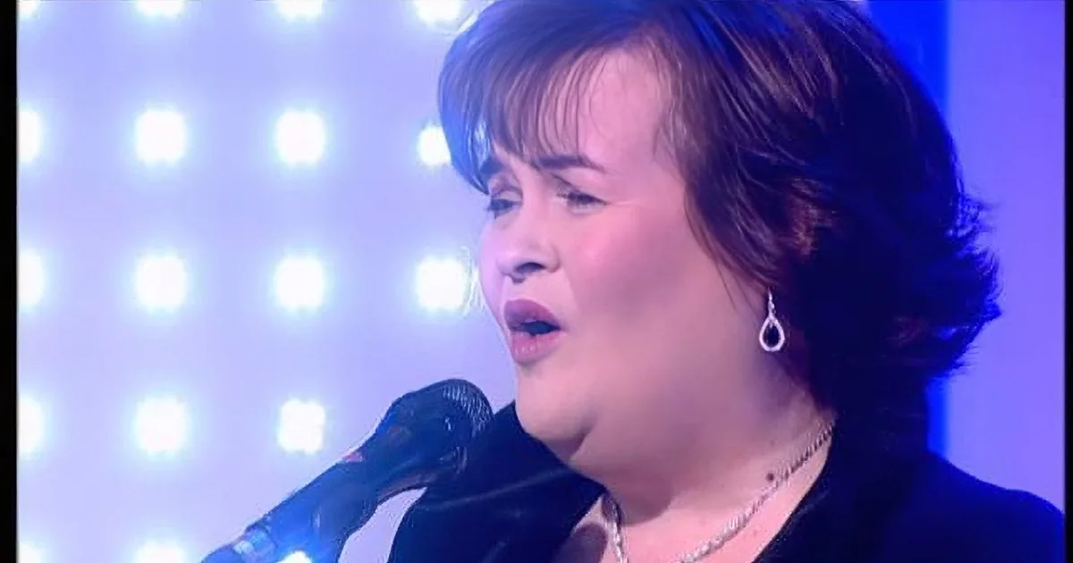 A woman with short hair, wearing earrings and a necklace, sings into a microphone against a backdrop of bright lights.