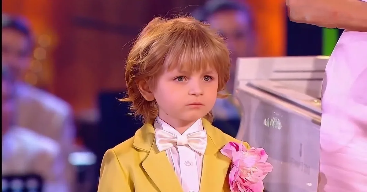 A young child in a yellow suit plays a white piano on a stage.
