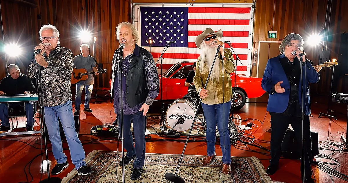 A group of men singing in front of an American flag backdrop with musical instruments and equipment around them.