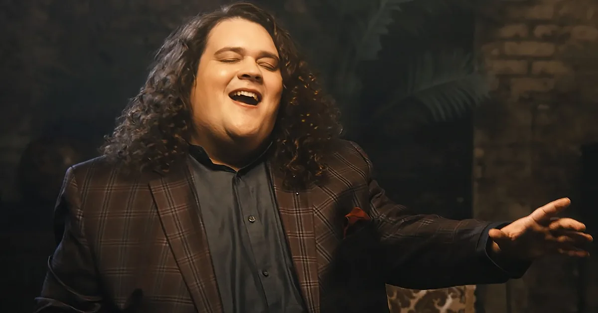 A man with long curly hair, wearing a plaid blazer and black shirt, extends his arms and smiles widely.