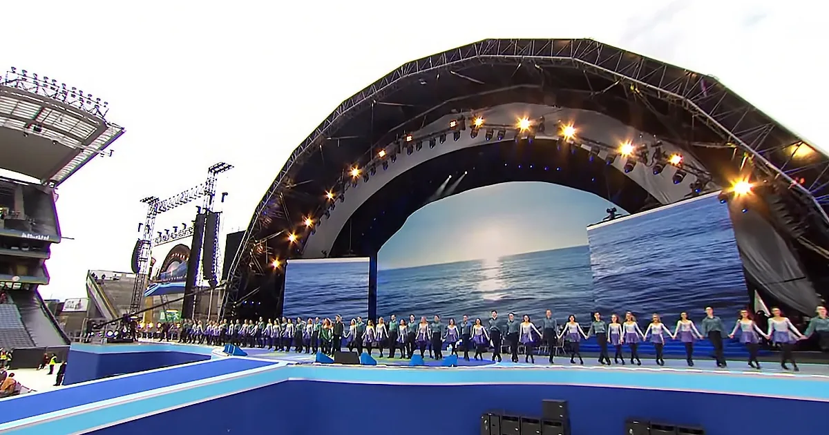 A large group of people in blue and white costumes stand in a line on a stage with a large screen displaying a sunset...