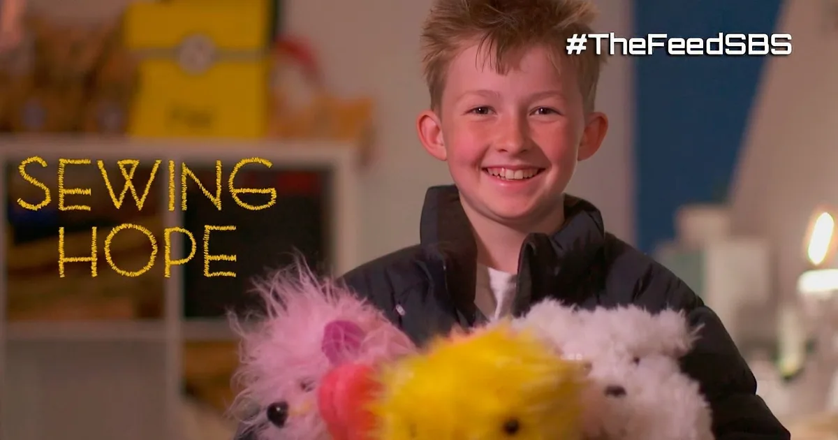 A boy smiles while holding plush toys in a room with sewing equipment and shelves.