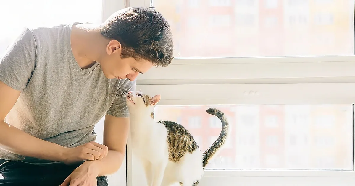 A man leans close to a cat by a window, both appearing calm and content.