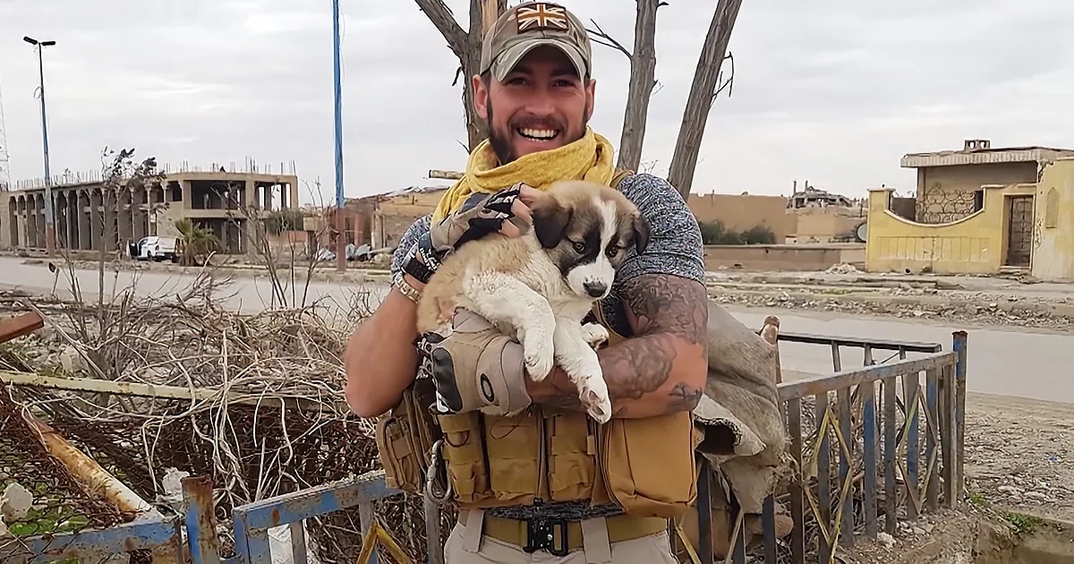 A man in military gear holds a puppy, smiling amidst a backdrop of damaged buildings and debris.