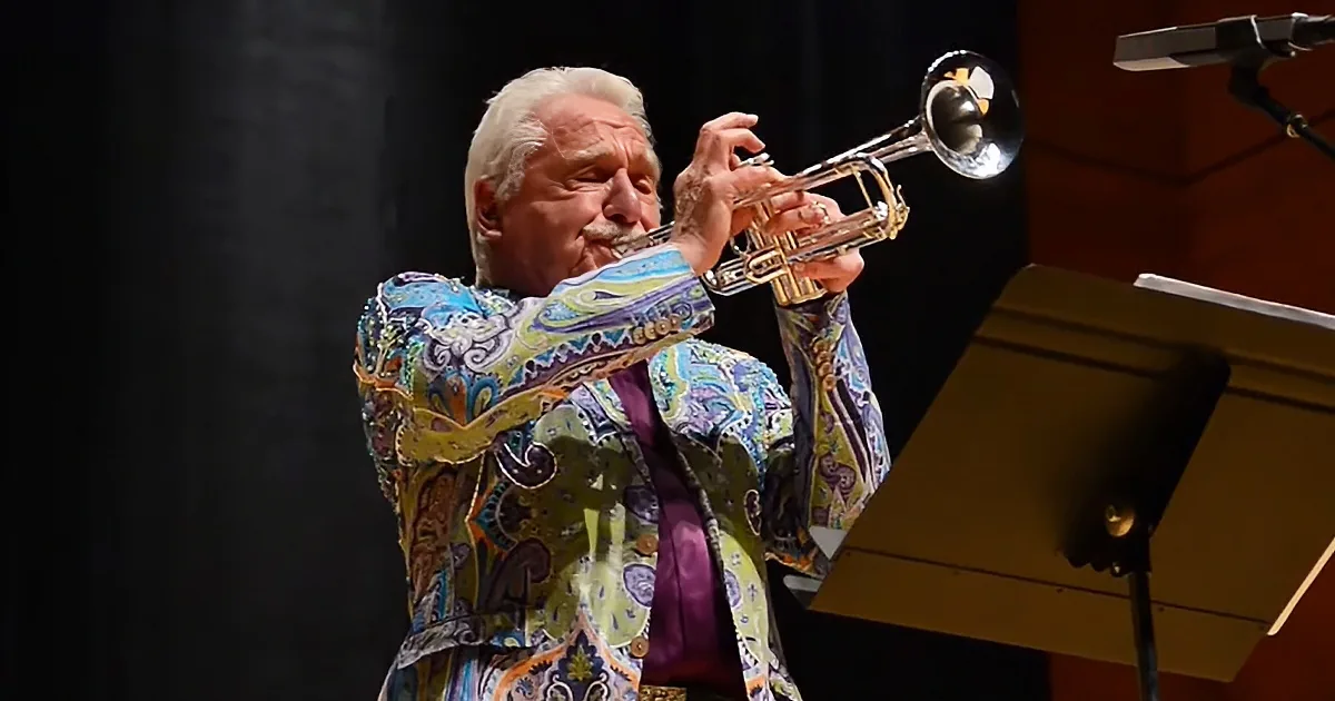 An elderly man in a colorful paisley jacket plays a trumpet on stage.
