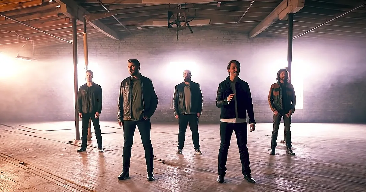 Five men in leather jackets stand in a dimly lit, smoky warehouse with brick walls and wooden beams.