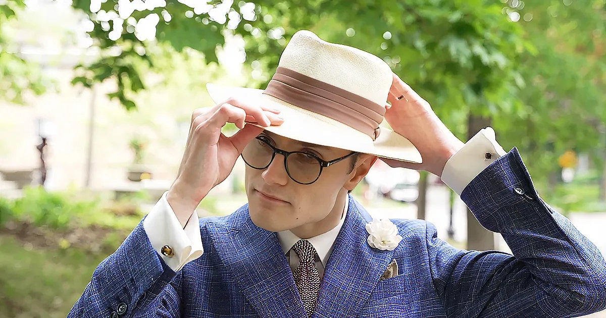A man in a blue plaid suit adjusts a beige fedora hat, wearing glasses and a white flower on his lapel.