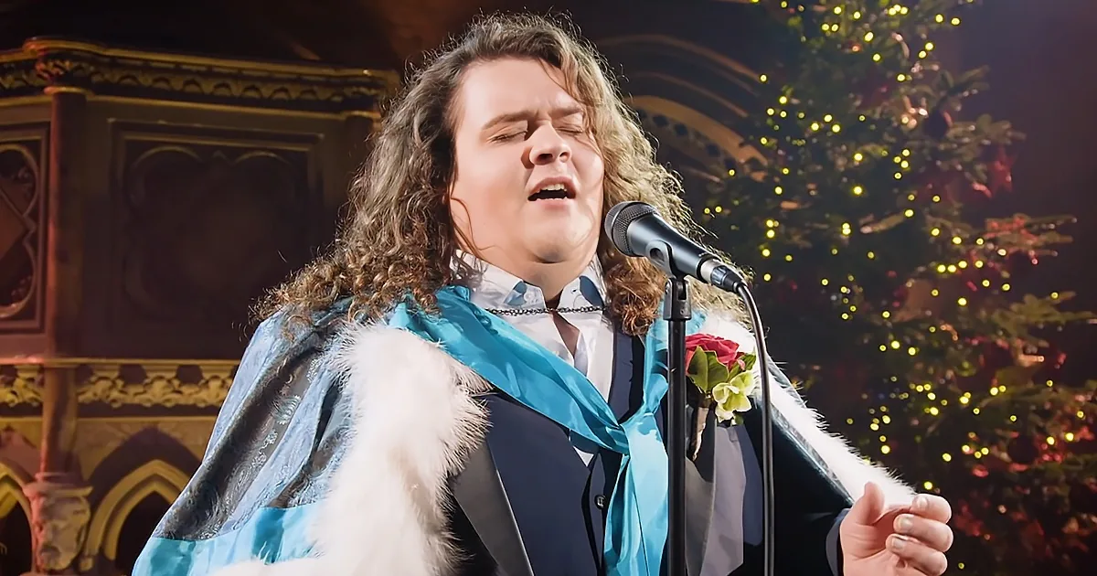 A person in a blue robe with a fur collar sings into a microphone, with a decorated Christmas tree in the background.