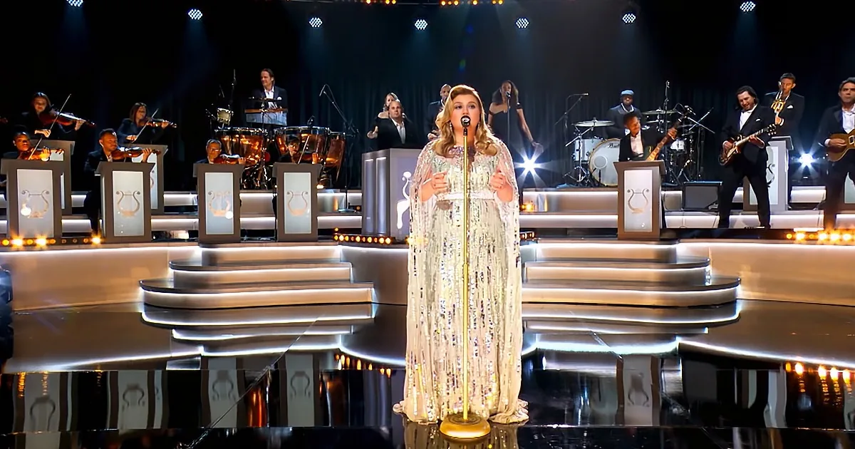A woman in a sparkling dress sings on stage with a band and orchestra behind her.