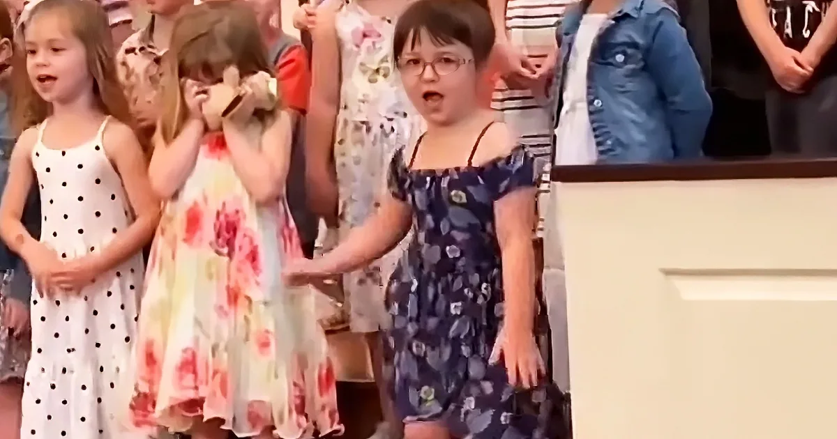 A young girl in a floral dress stands in front of a group of children and adults, singing with a microphone.